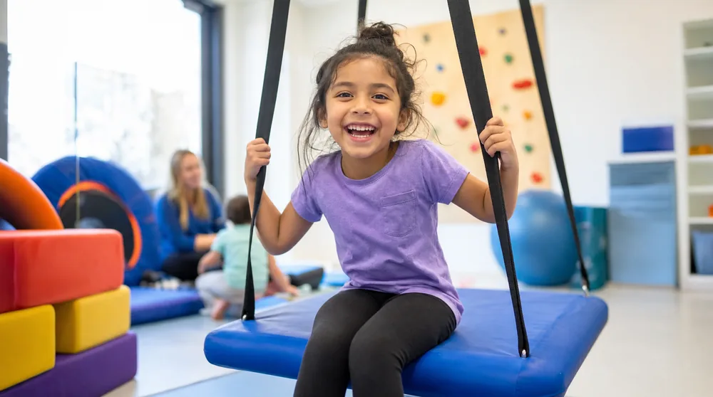 A joyful child swinging on a therapy sensory swing in a bright sensory gym